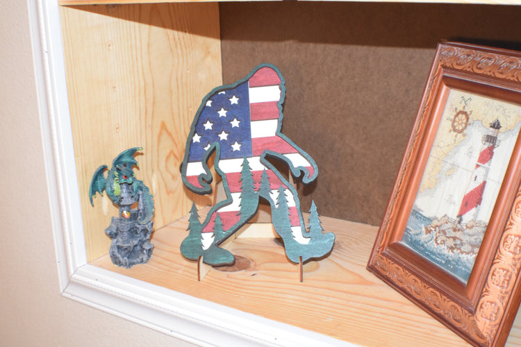 Patriotic Bigfoot. Layered wood art with the outline of Bigfoot with pine tree highlights and a background of the American flag.